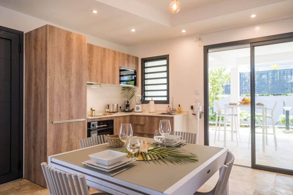 A modern kitchen with wooden finishes, featuring a grey table set for a meal with wine glasses, a plate, and plant decorations. In the background, a garden view is visible through large sliding glass doors.
