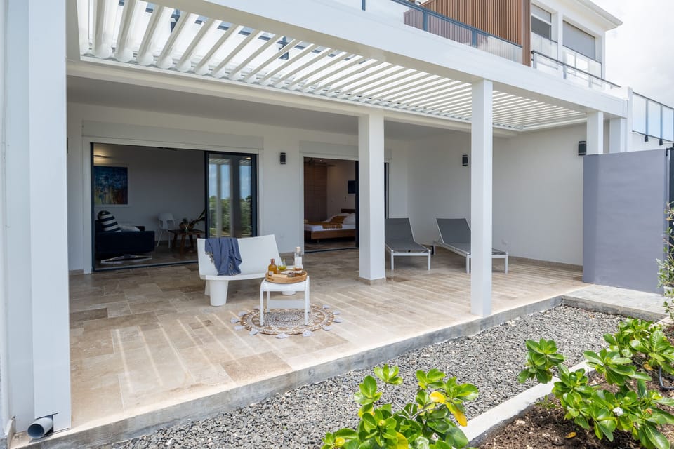 An elegant modern patio with white outdoor furniture, overlooking a bright interior room with artwork. Lush green plants add beauty to the outdoor space.