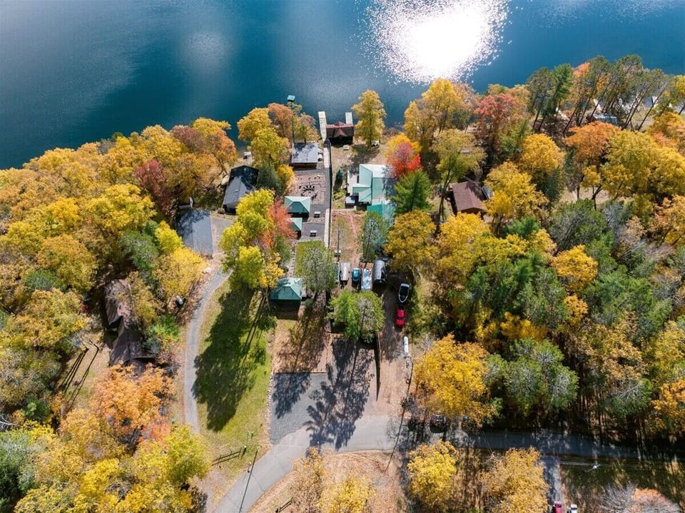 An aerial view of small, colorful cabins nestled in a dense forest of yellow and orange autumn trees along a blue lakeshore.