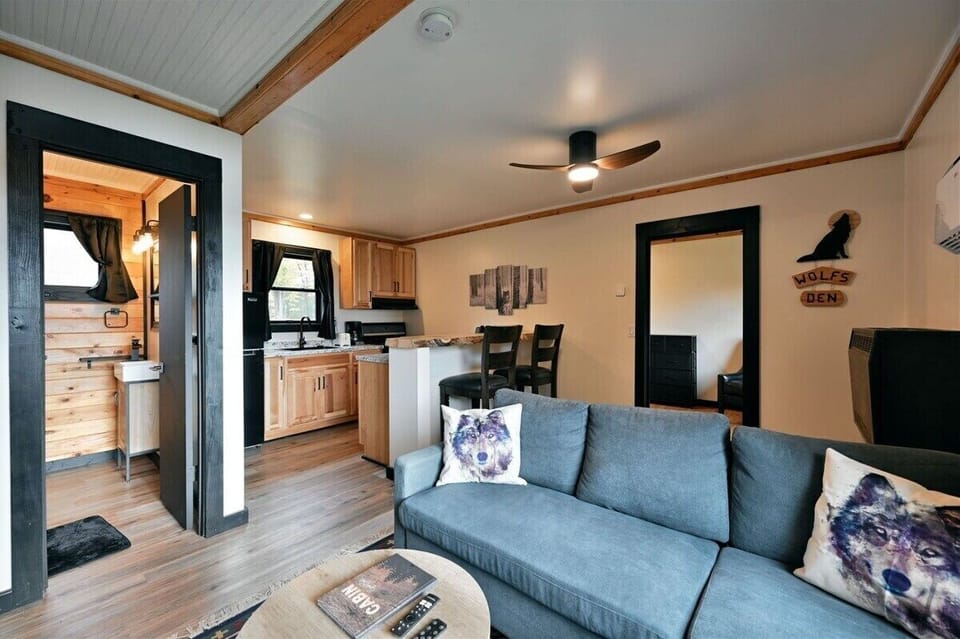 Interior of a modern cabin featuring a blue sectional sofa with wolf-print pillows, a wooden dining set, and a view into a light-wood kitchen.