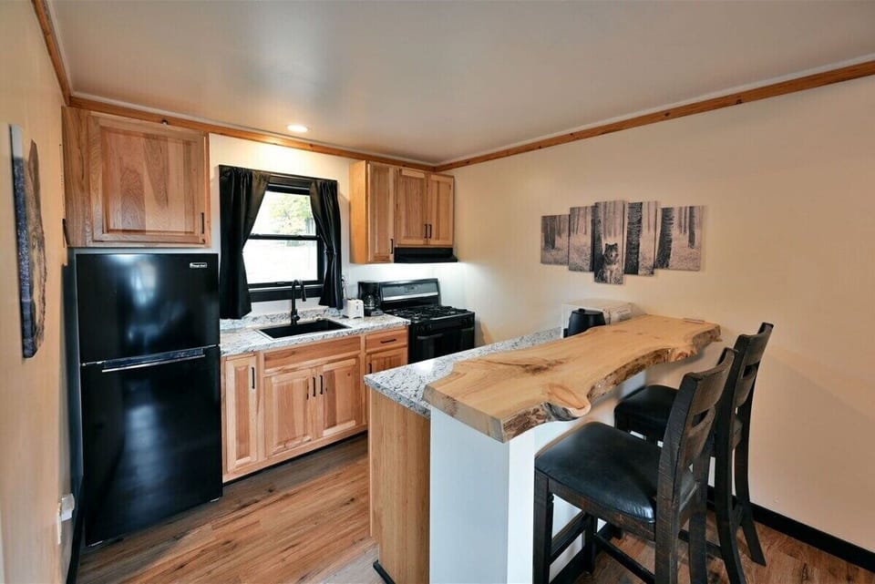 A compact kitchen with light wood cabinets, a black refrigerator and stove, and a live-edge wood breakfast bar with two black chairs.