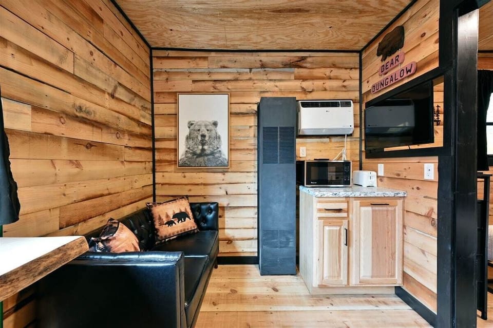 Living area of the Bear Bungalow featuring wood-plank walls, a black leather sofa with bear-themed pillows, a bear portrait, and a small kitchenette with a microwave and white cabinetry.