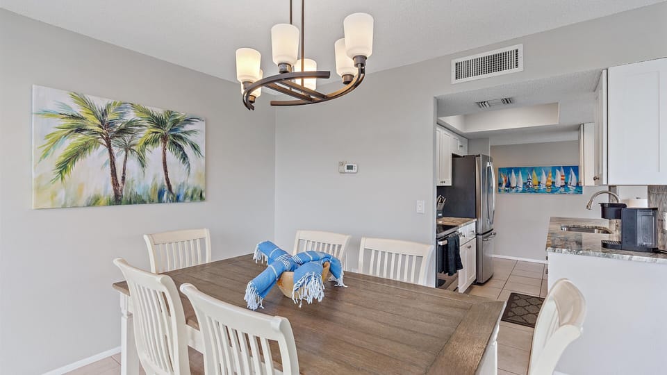 A dining area with a wooden table and six white chairs, a chandelier above, a wall painting of palm trees, and a view into a kitchen with stainless steel appliances and a sailboat-themed wall decoration.