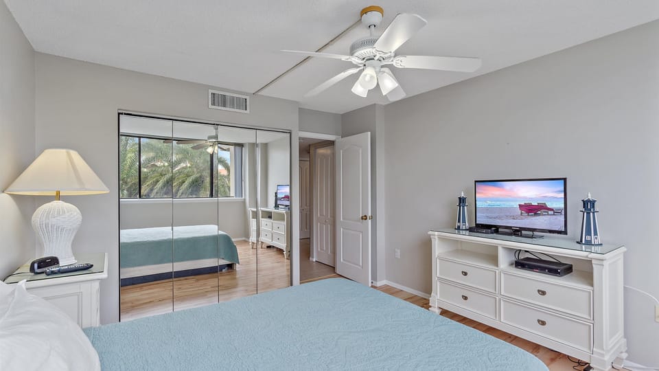 A bedroom with a ceiling fan, a TV on a dresser, a lamp on a nightstand, and a bed with a blue blanket. A mirrored closet and a window with blinds are on the left.