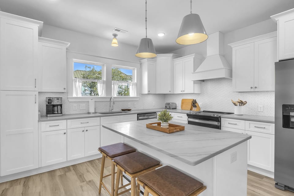 Bright coastal kitchen with white cabinets, island, 3 stools, pendant lights, stainless fridge, stove, vent hood, sink, coffee maker, toaster, tile backsplash, wood floors, and windows with tree views.