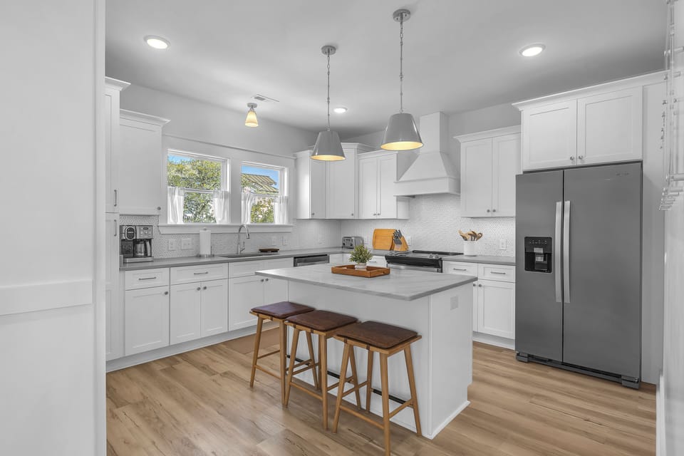 Bright coastal kitchen with white cabinets, island, 3 stools, pendant lights, stainless fridge, stove, vent hood, sink, coffee maker, toaster, tile backsplash, wood floors, and windows with tree views.