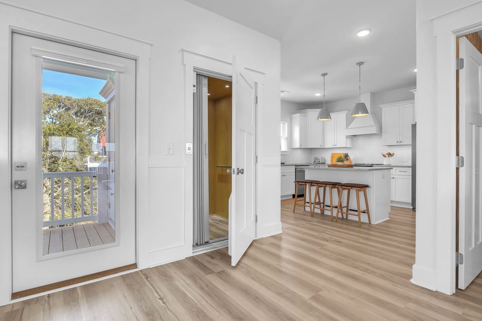 Bright coastal kitchen with white cabinets, island, 3 stools, pendant lights, stainless fridge, stove, vent hood, sink, coffee maker, toaster, tile backsplash, wood floors, and windows with tree views.