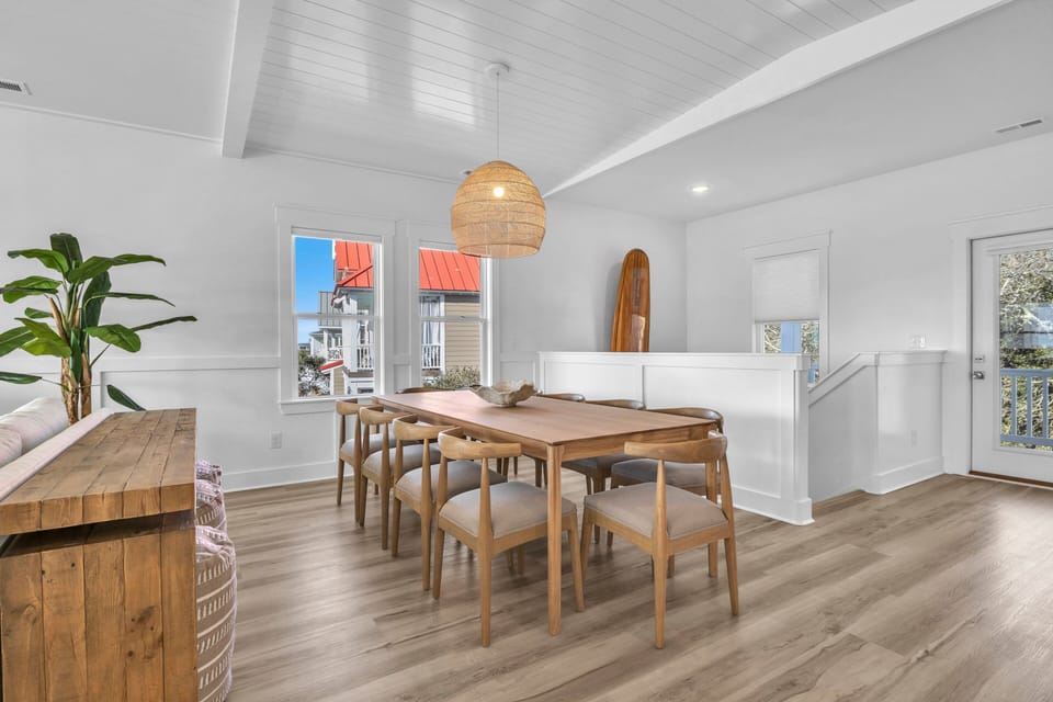 Bright dining area with wood table, 6 chairs, woven pendant light, plank floors, windows, stair rail, deck door, surfboard decor, plant, and console table with stools.