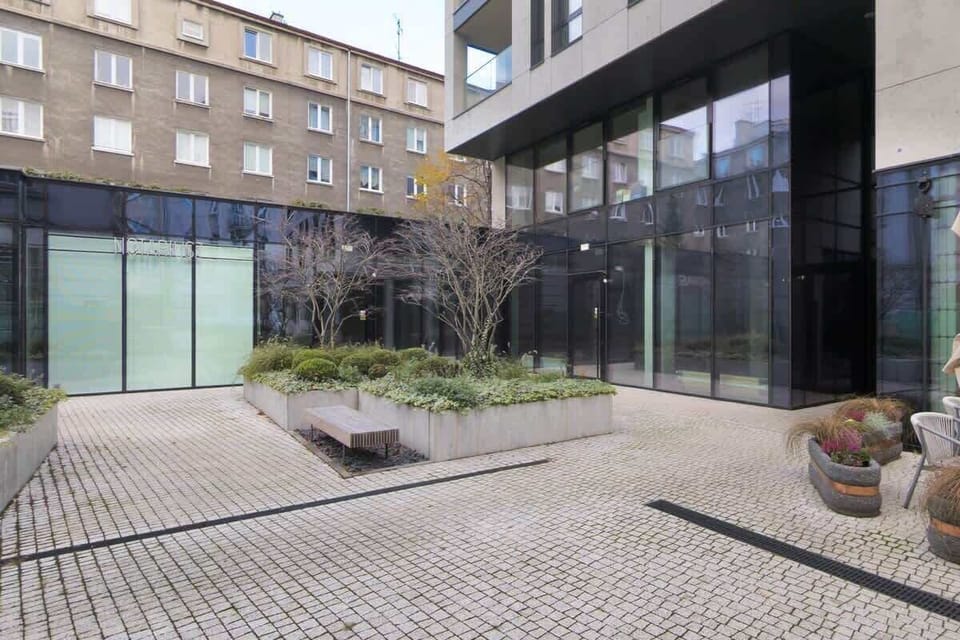 An outdoor courtyard with a paved walkway and open seating area, framed by contemporary building facades.

