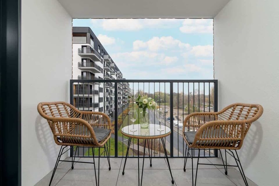 A cozy balcony setup with two rattan chairs and a small table, overlooking an urban view with blue skies and neighboring buildings.

