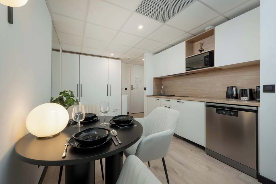 A chic dining area with a round table, and a modern kitchen in the background. The space has white cabinetry, sleek lighting fixtures, and is designed for functionality and style.