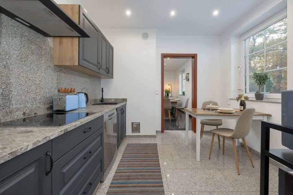 A modern kitchen with dark cabinetry, a speckled countertop, and open access to a dining table set against a windowed wall.

