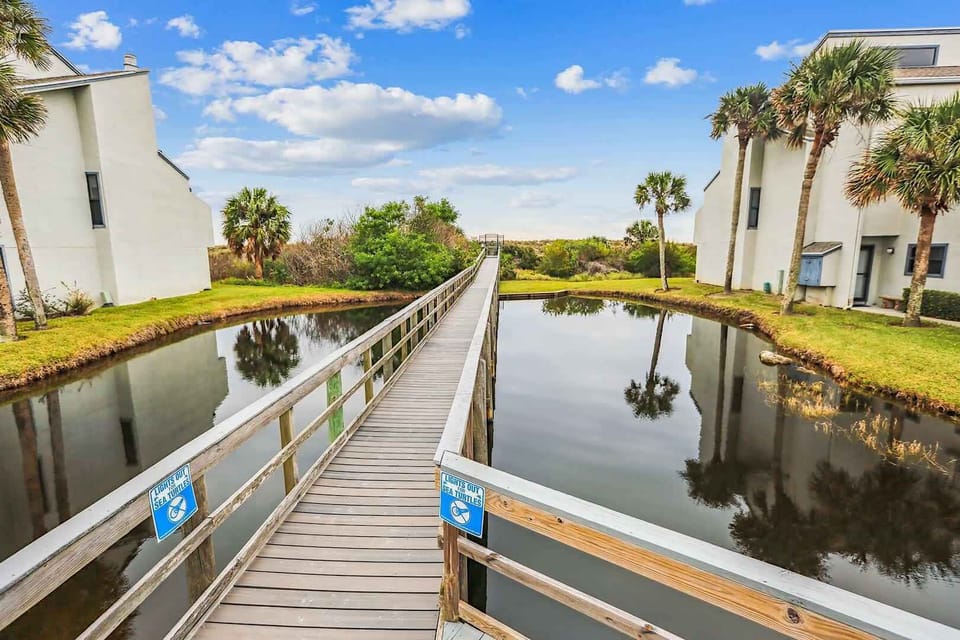 - Stroll across this charming boardwalk surrounded by calm waters, leading you toward coastal beauty.  
- Palm trees sway gently, framing peaceful reflections along the way.  
- A picture-perfect entry to your beach escape awaits every step.  
