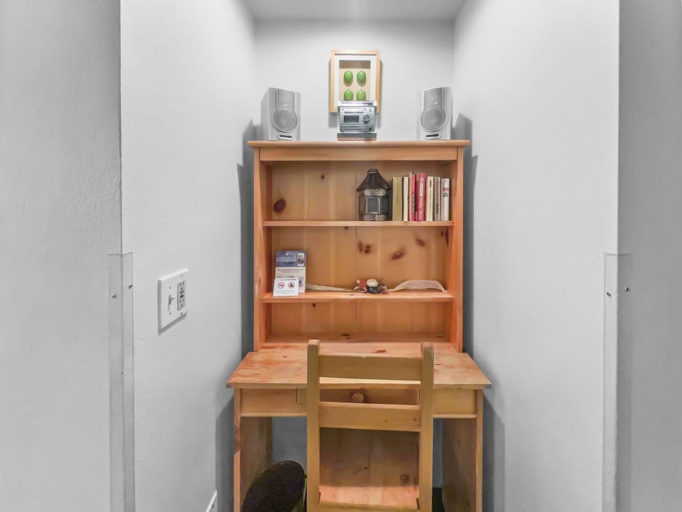A small wooden desk and chair are set in an alcove, featuring books, a small lamp, and decorative items on the shelves above. A framed picture and speakers are placed on the top shelf.