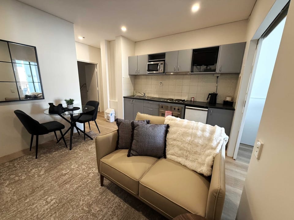 Relax in this open-plan studio featuring a cozy sofa, dining table for two, and a fully equipped kitchenette with a cooktop, oven, microwave, and fridge. Thoughtfully designed with modern furnishings and everything you need for a comfortable stay.