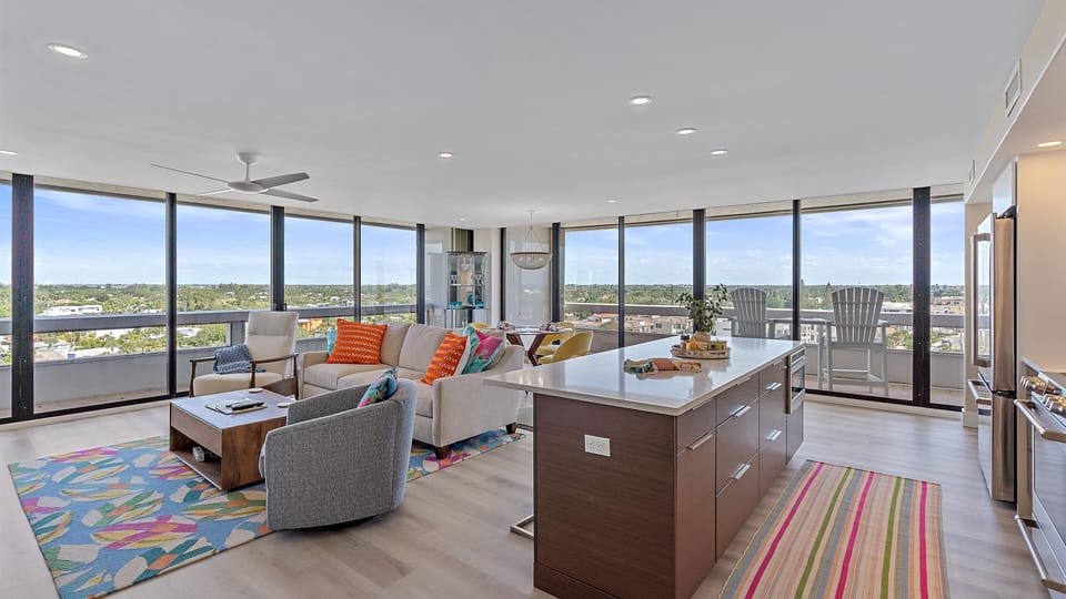Modern open-plan living room and kitchen with floor-to-ceiling windows, contemporary furniture, colorful rugs, and a cityscape view. The room is filled with natural light.