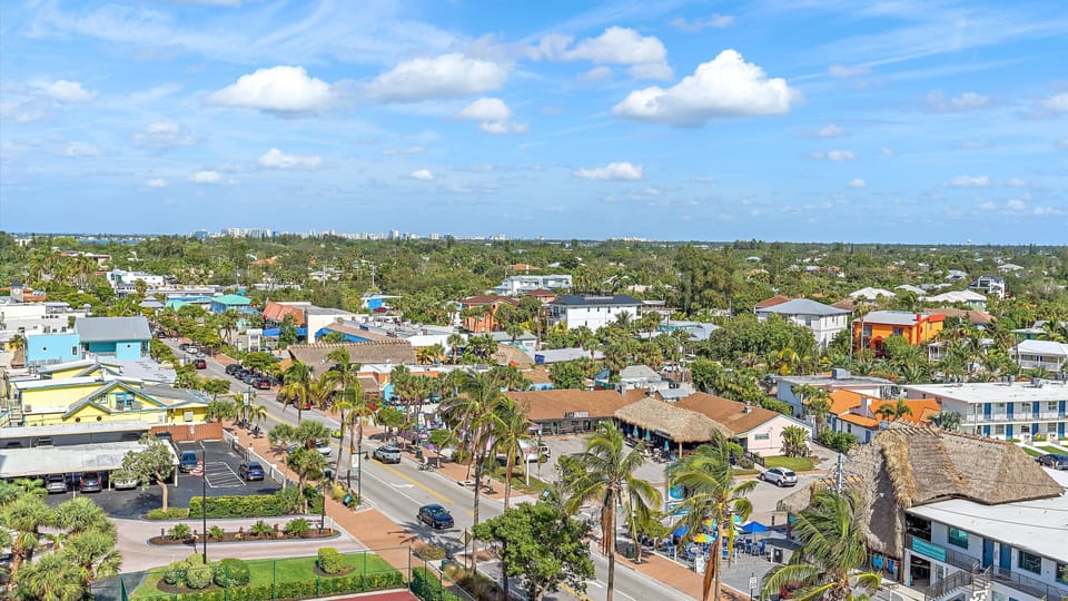 Aerial view of a suburban area with a mix of residential housing, commercial buildings, trees, and clear blue sky with scattered clouds.
