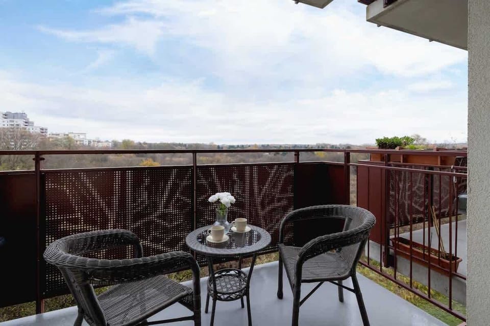 A balcony with a small round table and two wicker chairs, offering an outdoor relaxation spot with a view of the neighborhood.


