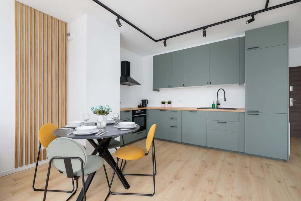 An open-plan kitchen with light green cabinetry, a round dining table with yellow chairs, and wooden wall accents for a cohesive, modern look.


