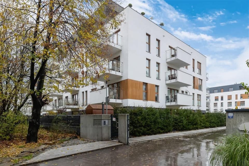 An exterior view of a modern residential building with white and wood-paneled facades, surrounded by well-kept greenery and a paved walkway.

