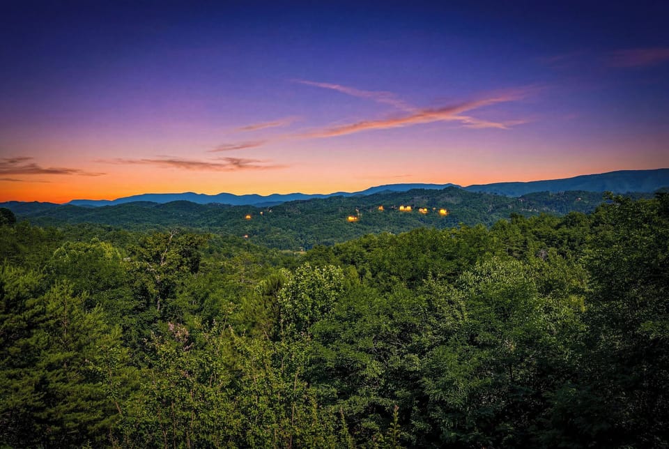 Stunning sunset over forested mountains and distant lit cabins, captured from the property's scenic surroundings—an idyllic natural backdrop for a mountain getaway.