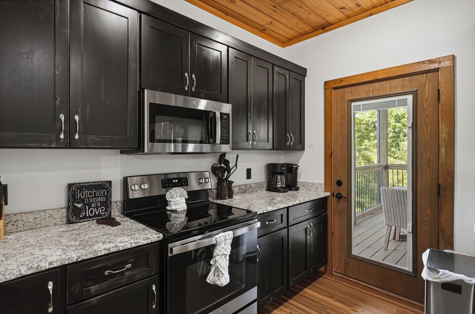 Stylish kitchen featuring sleek dark cabinetry, warm wood tones, granite countertops, and modern appliances. Wood-beamed ceiling adds rustic charm, while French doors open to your private deck.