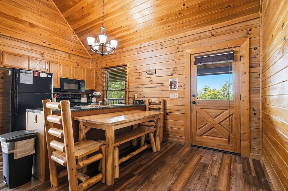 Step into your cozy cabin retreat with rich wood paneling and vaulted ceilings. Sunlight streams through, highlighting the inviting dining space and fully-equipped kitchen—your perfect home base.