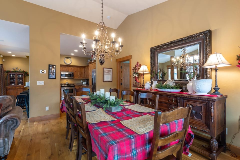 A dining room with a long wooden table covered in a red plaid tablecloth, surrounded by chairs, under a chandelier. A mirror and various decorative items are on the sideboard against the wall.