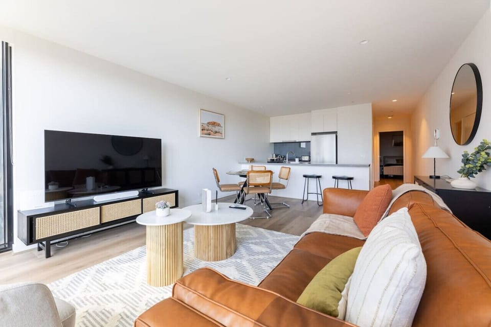 A bright, open-plan living space with a leather sofa, large smart TV, and a seamless flow into the dining area and kitchen. Warm natural tones and timber accents give the space a comfortable, lived-in feel.