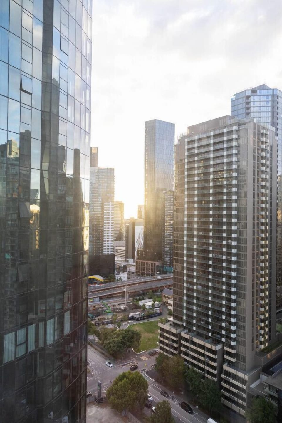 Golden sunlight breaking through the Melbourne CBD skyline, with glass towers, tree-lined streets, and city streets visible below — the kind of view guests can take in directly from the apartment.