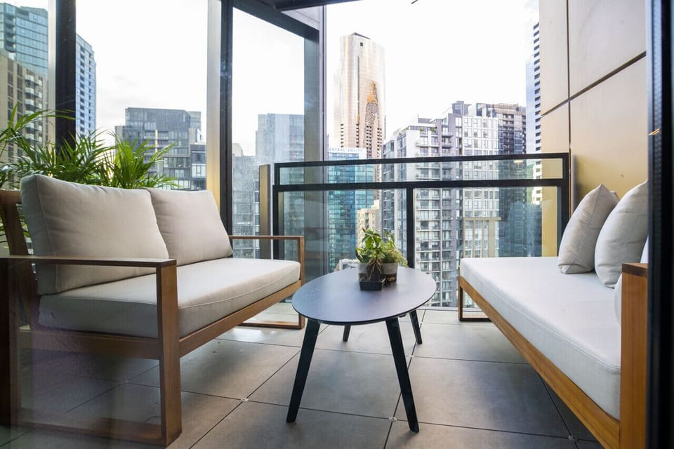 Private balcony with timber-framed outdoor seating, a round side table, and lush potted palms, all framed by sweeping glass balustrade views of the Melbourne city skyline.  *(171 characters)*
