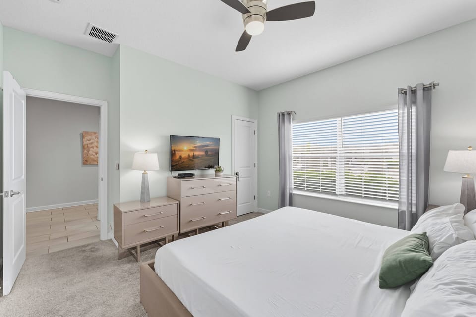 This bright bedroom features a soft green hue that complements the natural light streaming through the large window, while the plush bedding and stylish wood furniture create a cozy yet sophisticated atmosphere for relaxation.