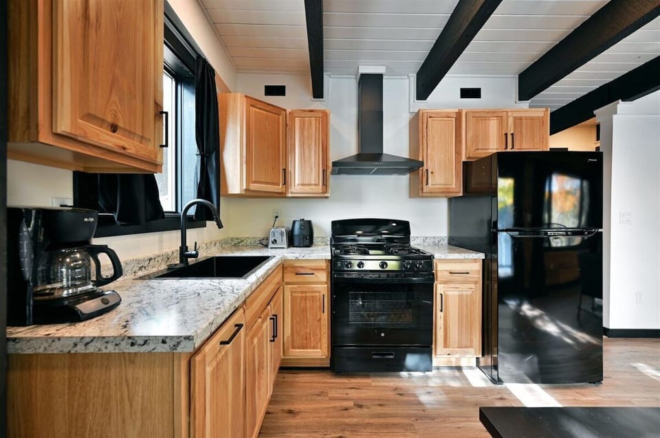 A compact kitchen in The Loon's Nest featuring light wood cabinetry, a black gas stove with a matching range hood, a black refrigerator, and a black farmhouse sink.