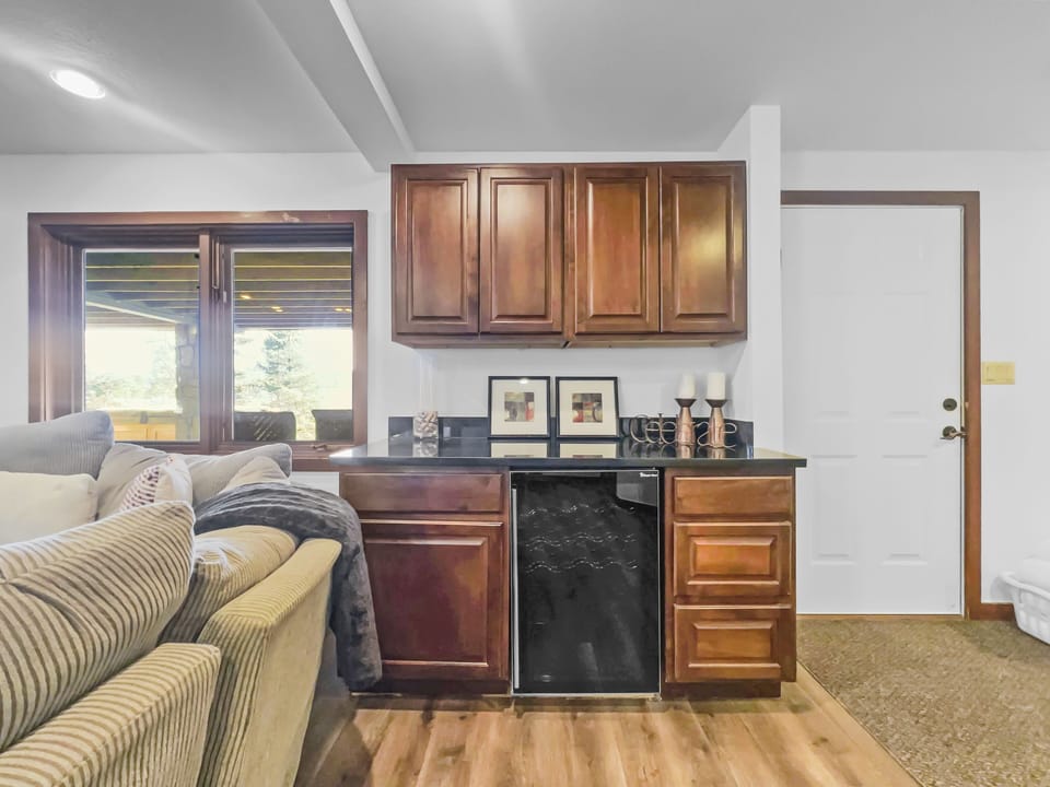 Small home bar area with wooden cabinets, a dark countertop, mini-fridge, artwork, candles, and a nearby sofa. Door and window are also visible.