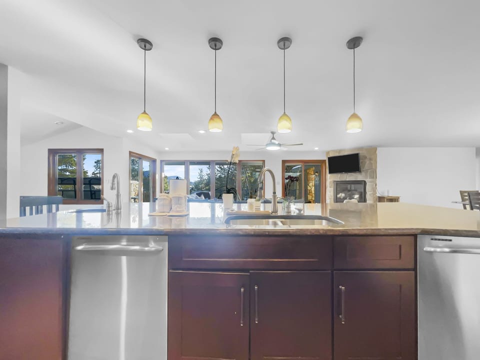 A modern kitchen with a large island sink, pendant lights above, stainless steel dishwasher, and wooden cabinets. Two sets of double doors open to a deck with seating. A fireplace is visible in the background.