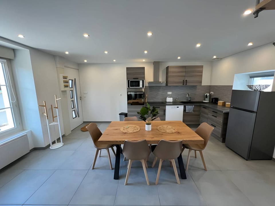 "This fitted and fully equipped kitchen is a friendly and functional space. The grey tiled floor creates an elegant contrast with the white walls, bringing a touch of modernity. In the center of the kitchen, a large wooden table seats up to six people on comfortable brown chairs."