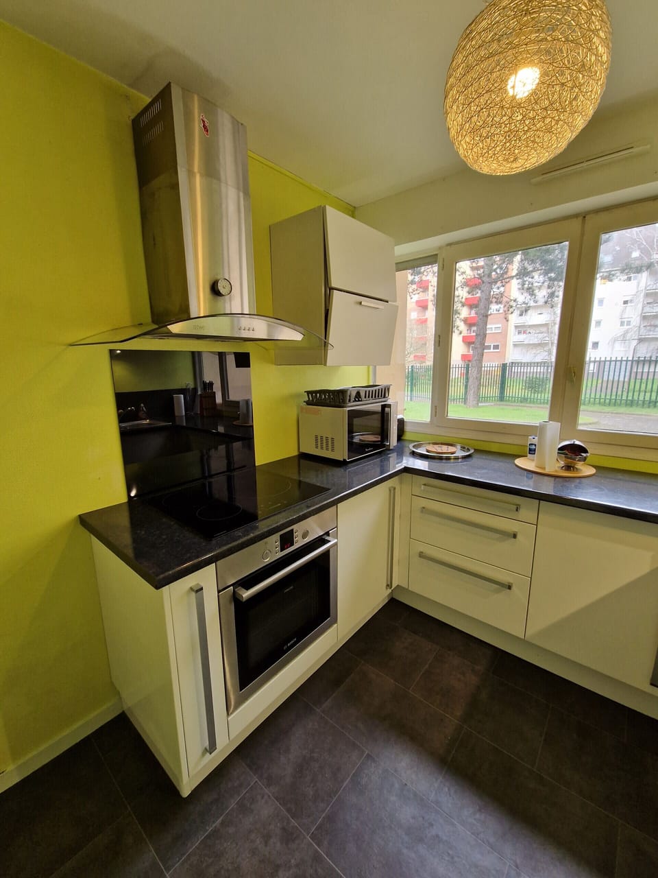 "Discover the chartreuse-yellow fully equipped kitchen of the Le Square property in Le Mans through this photo. The windows bring a beautiful luminosity to the space. You'll find induction hobs, a hood, a microwave and an oven for preparing delicious meals. The tall storage unit with its practical openings offers functional storage space. Enjoy a modern and aesthetic environment for your culinary activities."