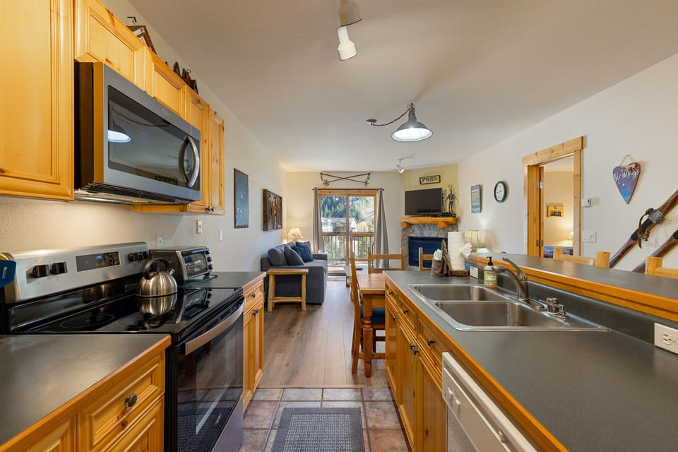 A modern kitchen with black countertops and wooden cabinets opens into a living area with a sofa, wall art, a mounted television, and a balcony with a forest view in the background.