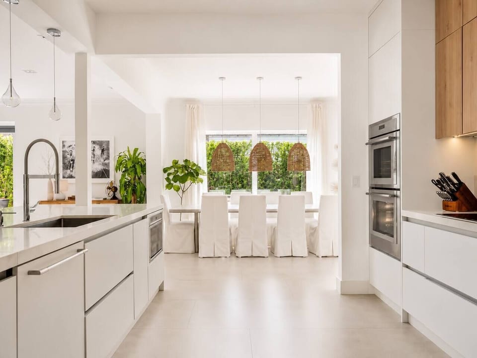Prepare meals in the well-stocked kitchen featuring a deep sink and white quartz island. The adjacent dining area, accented by three woven rattan pendant lights, offers a bright space for group breakfasts near the lush garden views.