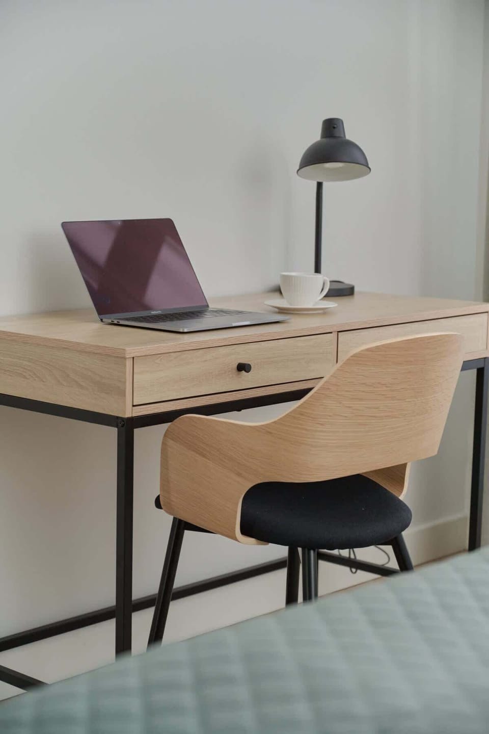 A minimalist workspace with a wooden desk, black chair, and a desk lamp placed next to the bed, suitable for remote work or study.
