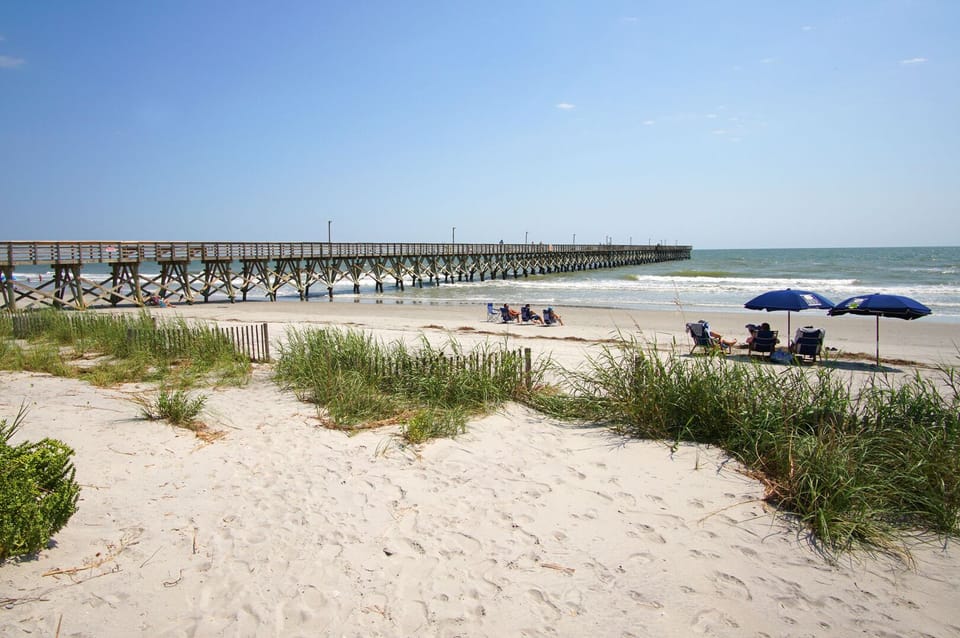 Beach with a pier, umbrellas and chairs steps from Sea Cabin 309, a 2-bedroom Cherry Grove rental