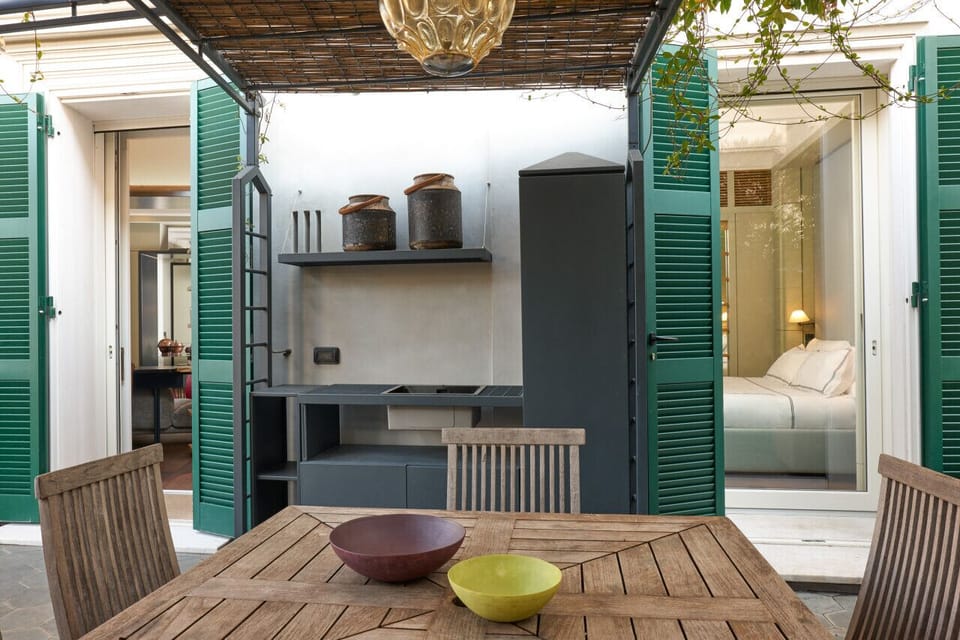 Outdoor area featuring a rustic wooden dining table and chairs, flanked by green shuttered doors leading to the interior. A sleek, built-in kitchenette with sink and shelving makes al fresco dining easy and stylish in this charming garden setting.