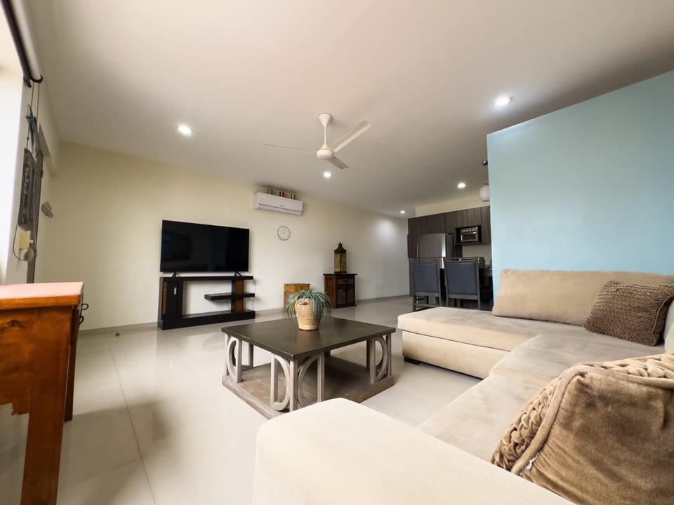 Living room with beige sectional sofa, square wooden coffee table, wall-mounted TV, and ceiling fan.