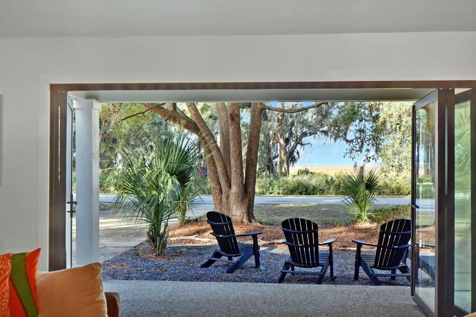View from Couch to Firepit - Open glass doors frame the front yard chairs, oak trees, marsh, and bridge beyond.
