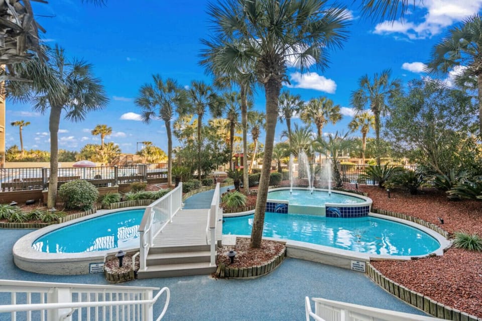 Tropical-style pool deck surrounded by swaying palms and glowing nighttime fountains. Unwind in the hot tubs, drift along the lazy river, and soak in the serene resort atmosphere beneath the stars.