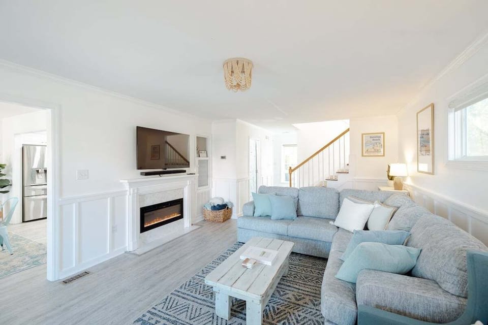 Bright and inviting living room featuring an electric fireplace, plush seating, blackout curtains, and coastal décor.