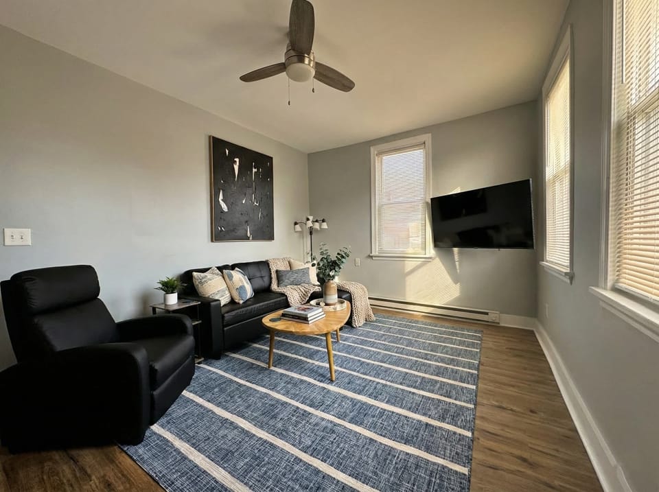 Living room with sofa seating, wall-mounted TV, ceiling fan, and large windows.