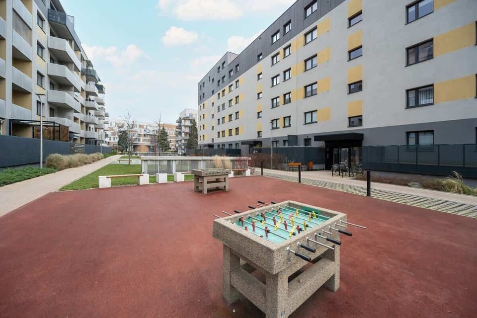 A recreational courtyard area featuring a foosball table, red rubber flooring, and surrounded by modern apartment buildings with yellow and grey facades.
