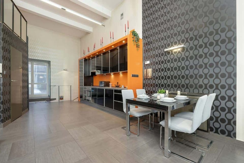 A modern dining area with a large table, elegant chairs, and orange-accented kitchen cabinets against a patterned wall.






