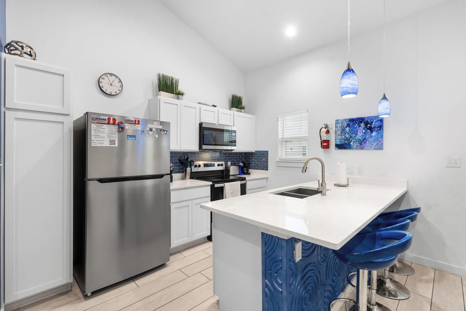 This modern kitchen invites you to gather around the sleek island with coastal-blue seating, where stainless steel appliances and crisp white cabinetry create an open, light-filled space perfect for preparing meals and making memories.